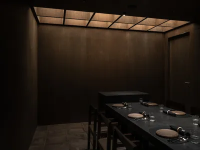 Dimly lit restaurant dining area with wooden tables, chairs, and wood paneling walls, featuring black place settings and a cozy atmosphere