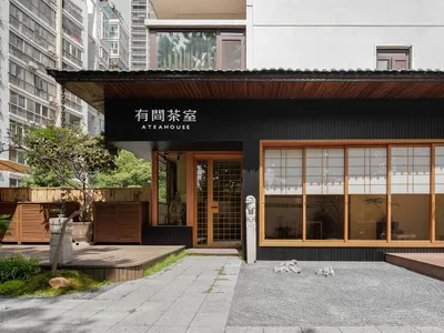 Modern Asian restaurant with a black facade, wooden elements, and natural light from windows, featuring a dining area with tables and chairs and potted plants
