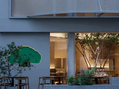 Modern restaurant interior with natural light, wood, and glass elements, multiple dining tables and chairs, and a warm atmosphere