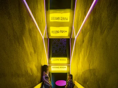Two women standing on a staircase in a dimly lit room with yellow lights, chairs, and a mix of living and hallway space