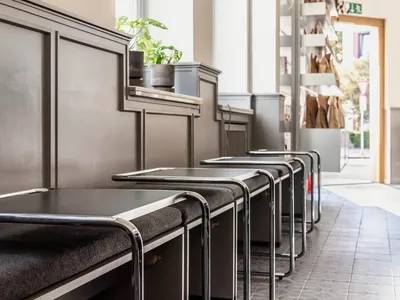 Modern restaurant interior with a long dining table and metal chairs, surrounded by potted plants and natural light