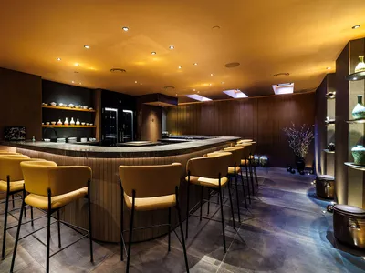 A modern restaurant with a large bar area featuring a wood countertop and metal stools, surrounded by chairs and tables in a warm and inviting atmosphere