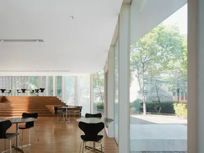 Dining area with multiple tables and chairs, glass walls, and wooden flooring, allowing natural light to enter the space