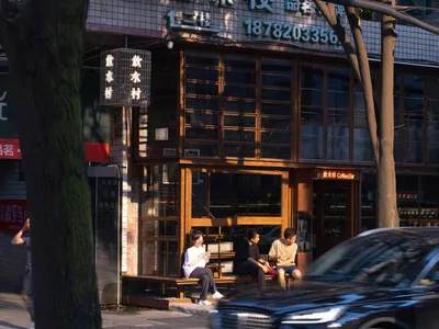 An Asian restaurant with people sitting outside on wooden benches in front of the establishment, surrounded by glass walls and natural light