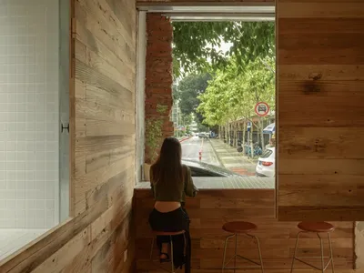 A woman sits on a stool at a counter in a cozy living space with wood flooring, earth tones, and a potted plant, surrounded by three chairs