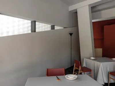 A restaurant dining area with two tables set with red chairs, white tablecloths, and tableware, with a kitchen visible in the background