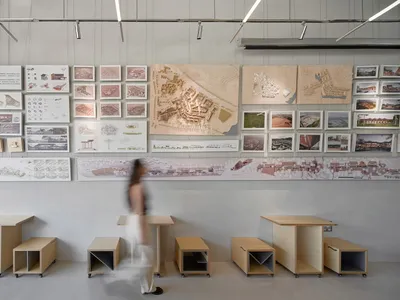 室內設計展廳牆壁與桌上陳列建築圖面，一位女性穿梭於展場空間
