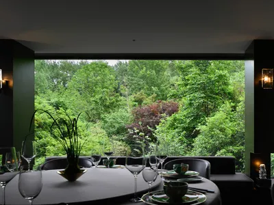 Dining area with a large window offering a view of trees, elegant table setting with wine glasses, bowls, and cups, and comfortable seating