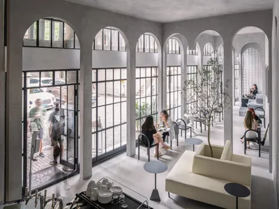 Spacious living area with a couch, chairs, dining table, and potted plants, set in a large white building with multiple windows and arches