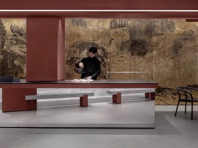 A man prepares food at a wooden counter in an Asian restaurant with a red wall and two chairs
