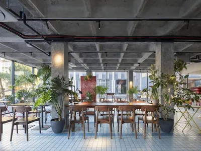 Modern restaurant interior with a large dining table, red accent, and potted plants, surrounded by chairs in a warm and inviting atmosphere