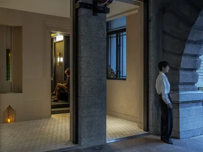 A hotel apartment hallway with a tiled floor, multiple doors, and two people waiting near their rooms