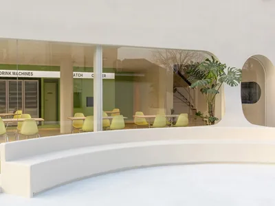 A modern restaurant with comfortable seating areas, natural light, and potted plants, featuring various chairs and benches around dining tables