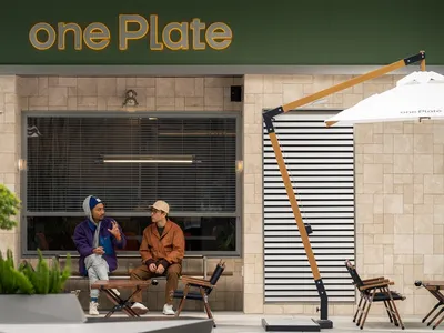 Two men sit in a modern cafe with a storefront window, surrounded by wood and glass elements, potted plants, and a casual atmosphere