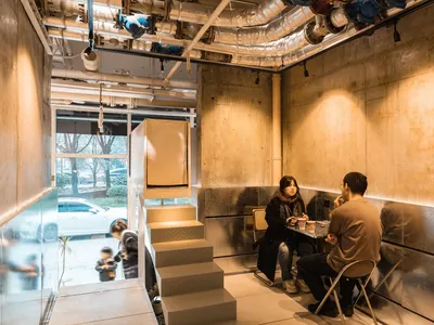 A casual restaurant dining area with concrete walls, chairs, and tables, featuring patrons enjoying meals and conversation