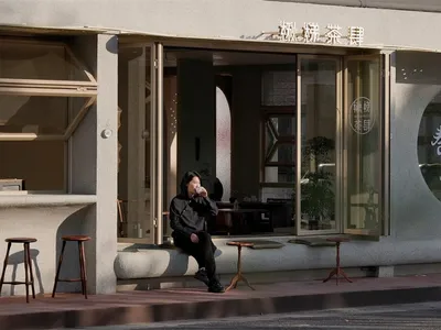 A woman sits on a bench outside an Asian restaurant, surrounded by chairs and potted plants, enjoying a cup of something in a relaxed and inviting atmosphere