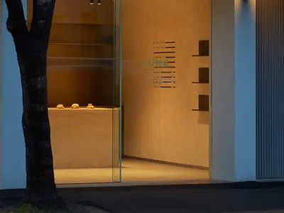 A brightly lit storefront with a glass door, surrounded by two trees and featuring three cakes on display