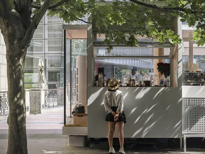 A casual outdoor cafe scene with a woman standing in front of a small white food stand, surrounded by potted plants and other people in the background