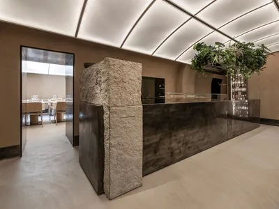 A restaurant interior with a stone wall separating two dining areas, featuring chairs, tables, and potted plants in a relaxed atmosphere