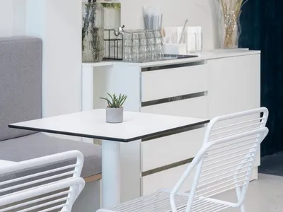 A clean and organized kitchen with a dining table and chairs, surrounded by white surfaces and a vase with a plant on the countertop