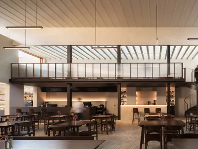 A modern restaurant interior with a large dining area featuring wooden floors, white walls, and well-lit tables and chairs, with patrons seated and standing