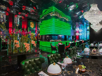 A modern restaurant with a large dining area featuring multiple tables, chairs, wine glasses, cups, and bowls, set against a backdrop of green walls