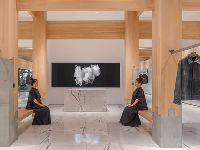 Two women sit on benches in front of a display wall featuring a large artwork, set against a backdrop of marble flooring and sophisticated decor