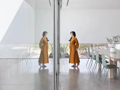 Two women standing in front of a glass wall in a dining room with chairs and a potted plant, reflected in the mirrored surface