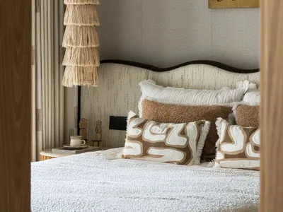 Cozy bedroom with a white bed, brown pillows, a wicker lamp, and a standard lamp, a chair, and a dining table