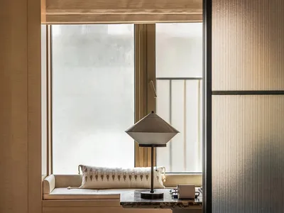 Cozy hotel living area with a couch, table, lamp, and bed in the background, featuring Asian-inspired decor and natural light
