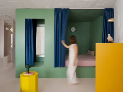 A woman walks through a calm and inviting bedroom in a green house with a bed, chairs, and a book, surrounded by white furniture and a serene atmosphere