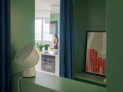A woman stands in front of a double sink in a green kitchen with a refrigerator and oven, surrounded by a cozy atmosphere