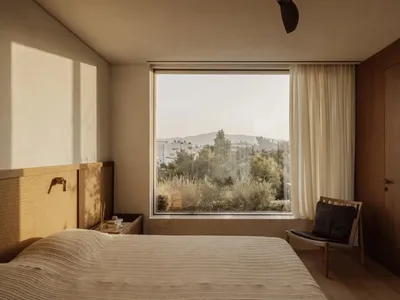 Cozy bedroom with a large window offering a city view, featuring a bed, chair, and two couches, with natural light creating a warm ambiance