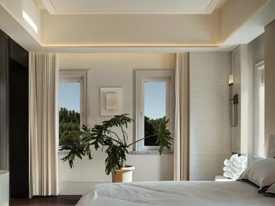 A bright bedroom with a large window, two smaller windows, and a bed extending from one end of the room to the other, with a potted plant near the foot of the bed