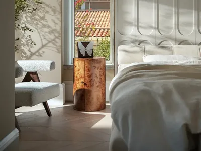 Well-lit bedroom with a white bed, wooden chair, potted plants, and a vase, set against a white wall