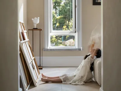 A woman sits on a window seat in a room with a white wall, looking out at a car parked outside through a large window, with two vases placed nearby