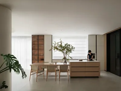 Modern kitchen with wooden cabinets, white walls, and a large window allowing natural light to fill the space, featuring two dining tables and several chairs, with a potted plant adding greenery