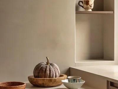 A kitchen countertop with three bowls, two cups, two spoons, and a pumpkin, set against a neutral background