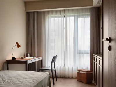 A bedroom with a bed and desk, natural light from two windows, and a lamp, set against a clean and bright white wall background