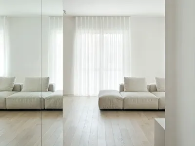 Empty living room with white walls, wooden floors, two couches, a chair, and natural light from windows