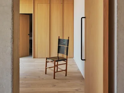A cozy living area with a wooden floor and a concrete wall, featuring a chair and a backpack, set against a neutral background
