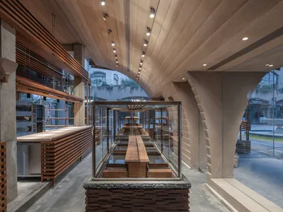 A modern restaurant interior with a long wooden table, chairs, and a counter with sink and refrigerator, featuring warm and inviting lighting