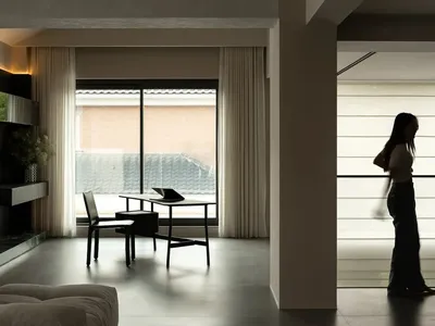 Woman walking towards a dining table in a modern living room with a couch, lamp, and white and grey colors, dim lighting creating a cozy atmosphere