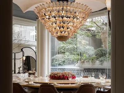 Elegant dining room with a semi-circular seating arrangement, a large chandelier, and potted plants, set for a meal with wine glasses, forks, knives, and spoons