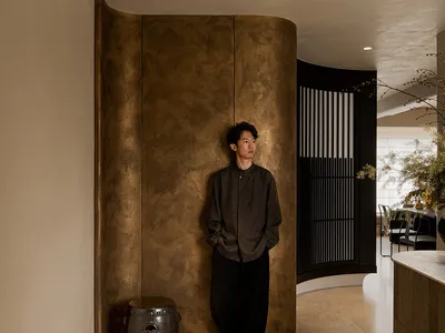 A calm and inviting living space with a brown wall, a dining table, and a chair, with a man standing in the center of the room