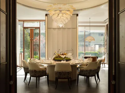 A well-lit dining room with a round table, chairs, and a chandelier, set amidst a larger living space with a couch and potted plant