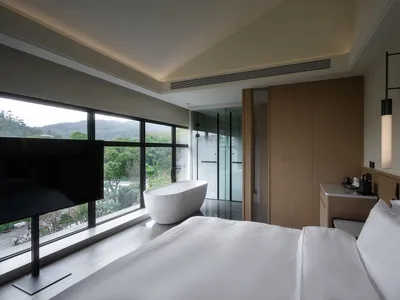 A well-lit hotel bedroom with a bed, bathtub, and wooden door, featuring a large window with a view of the outdoors