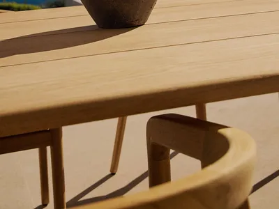 A wooden dining table with a large brown vase on a patio overlooking the ocean, surrounded by chairs in a serene and relaxing environment