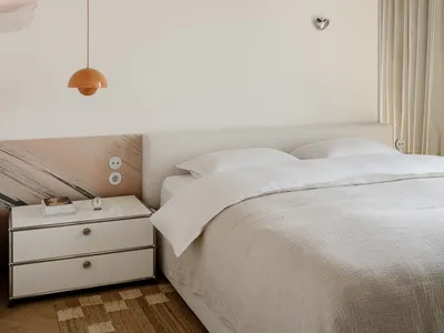 A minimalist bedroom with a white bed, dresser, and lamp, well-lit by natural light through the window