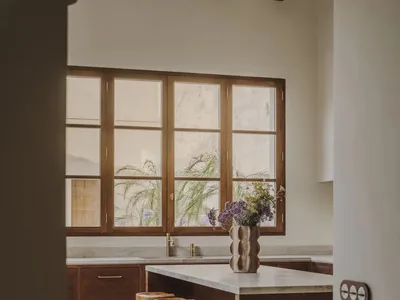 A modern kitchen with a wooden countertop, sink, refrigerator, and stools, featuring a vase with flowers and a window, with soft and warm lighting